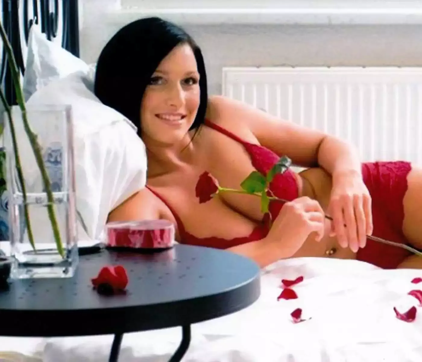 Eine Frau in sexy roter Unterwäsche und einer Rose in der Hand liegt lächelnd auf einem Bett